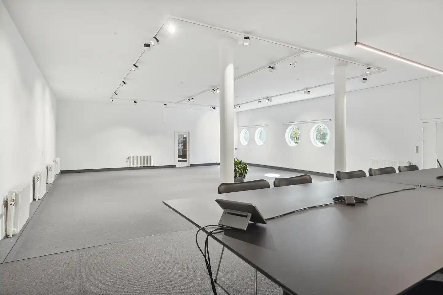 En tom mødehal med lysegult gulv, to lange mødeborde og høje lysekopper i loftet