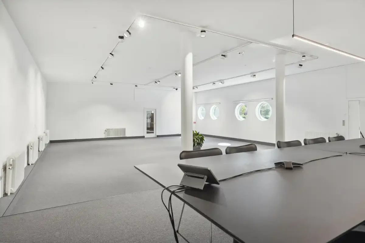 En tom mødehal med lysegult gulv, to lange mødeborde og høje lysekopper i loftet