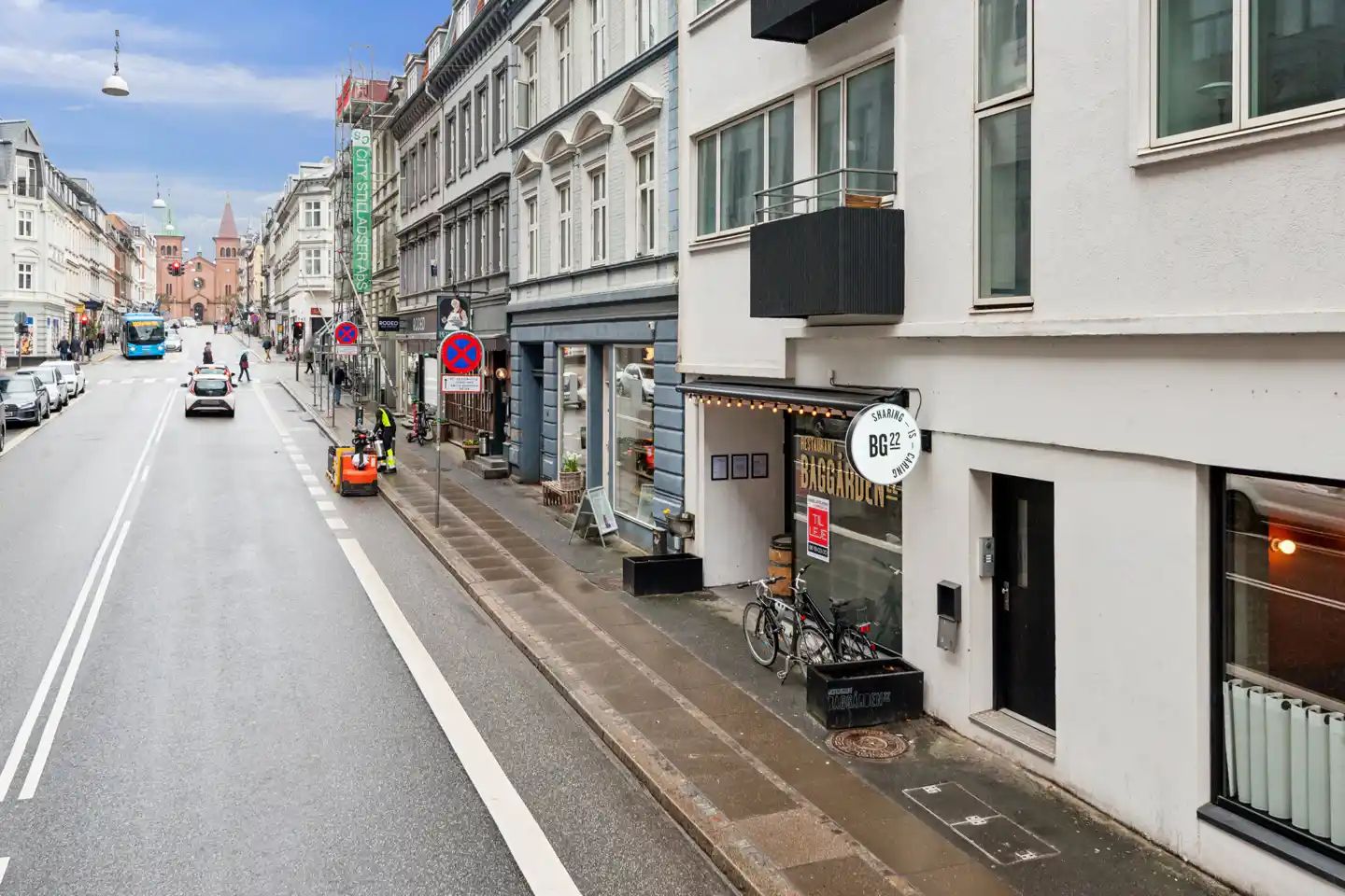 En bil kører ad en tosporet gade med restauranter og butikker. Mærkeplader, søjler og cykler er synlige langs siderne.
