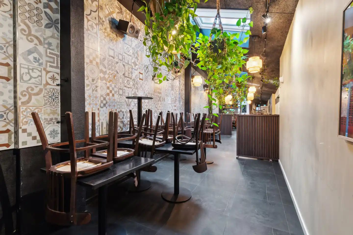Inde i en restaurant med tom bord, hvor stole er skubbet sammen. Væggene er dækket med mønstrede fliser og hængende planter.