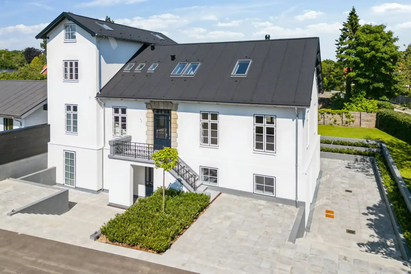 Bygning med terrasse, balkon og trappe, smykket med grønne anlæg og parkerede biler.