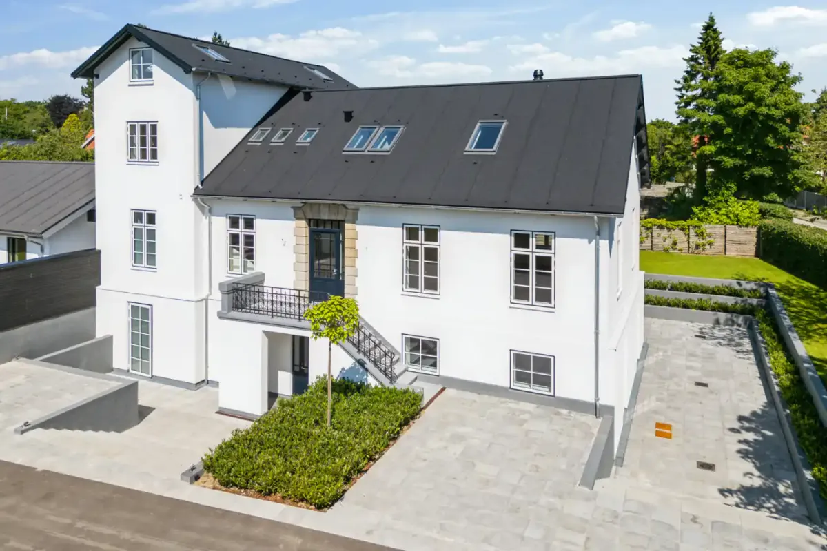 Bygning med terrasse, balkon og trappe, smykket med grønne anlæg og parkerede biler.