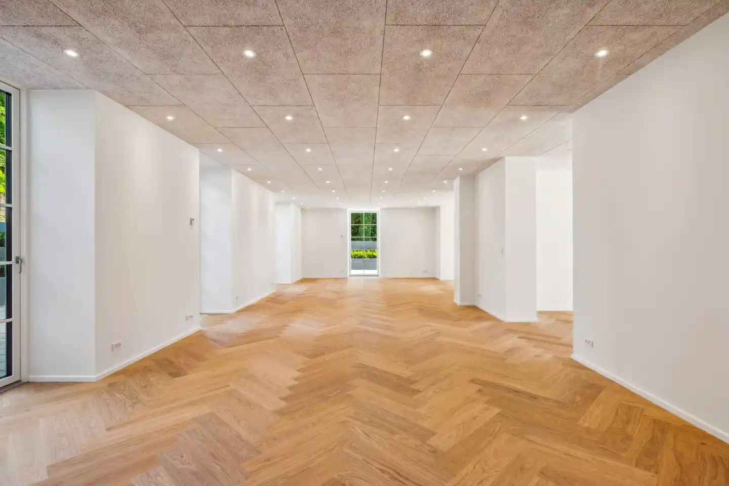 En stor, lyse, tom rum med en hvid væg og trægulvet. To vinduer i højre side og en dør til venstre.
