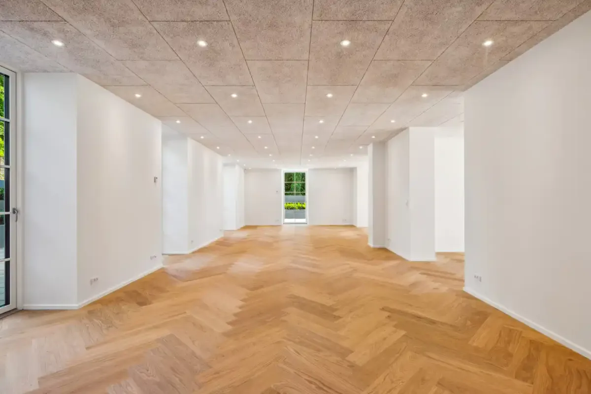 En stor, lyse, tom rum med en hvid væg og trægulvet. To vinduer i højre side og en dør til venstre.