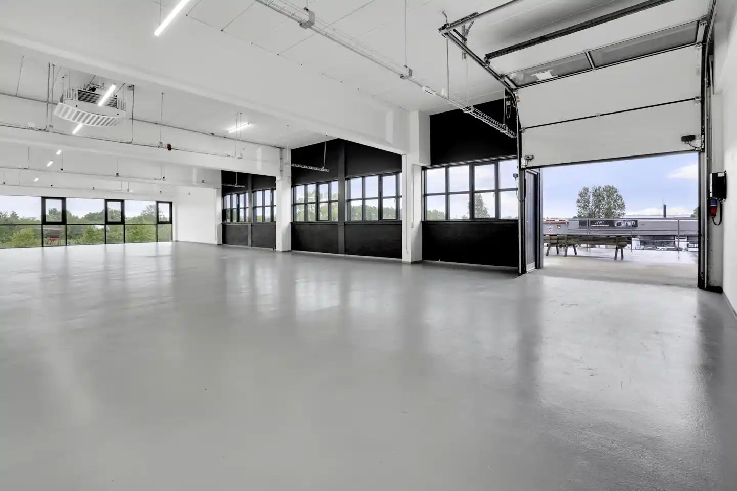 En stor rummelig kontor/showroom med hvide vægge, glasvinduer og højloft. Der er store garagedøre og en