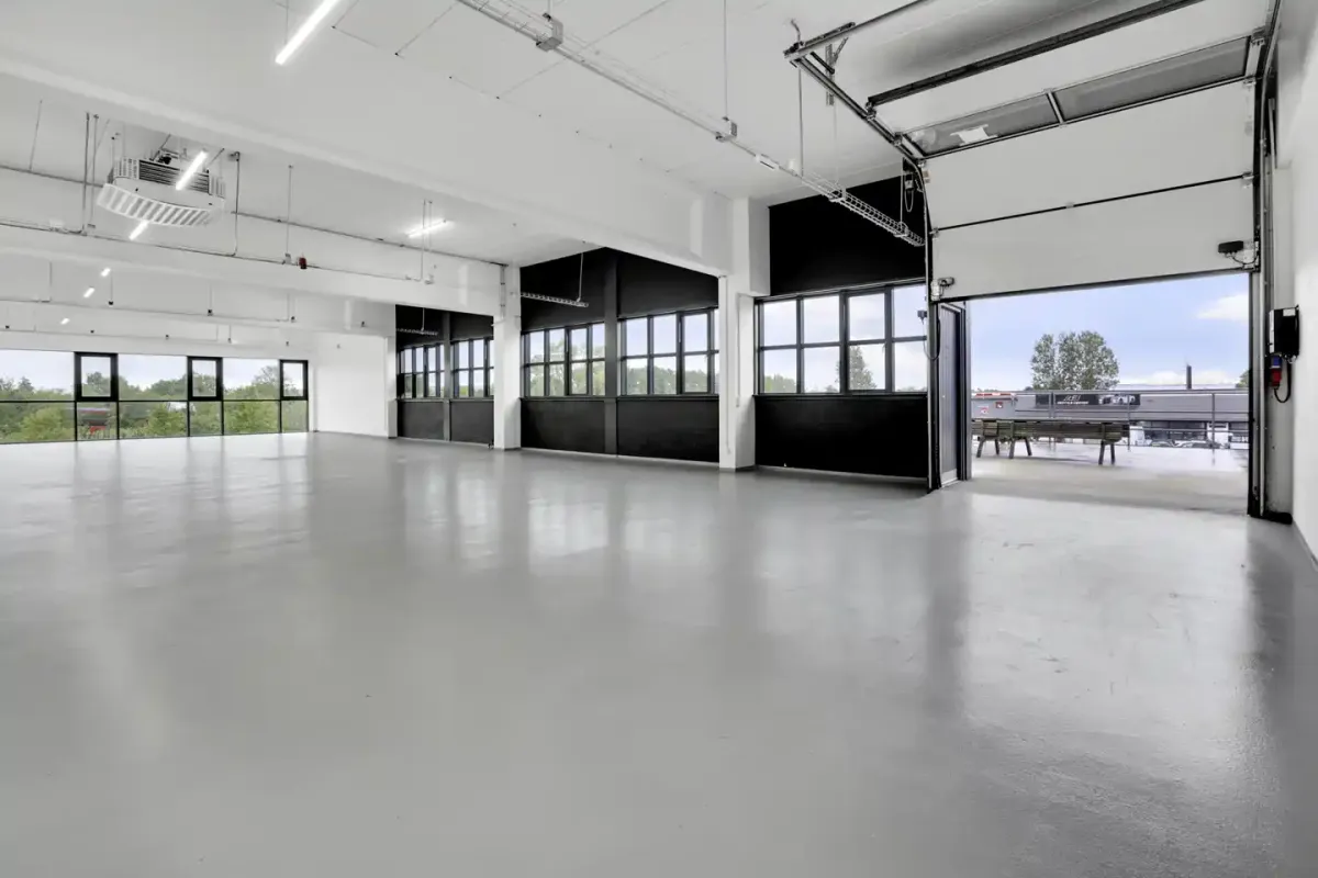 En stor rummelig kontor/showroom med hvide vægge, glasvinduer og højloft. Der er store garagedøre og en