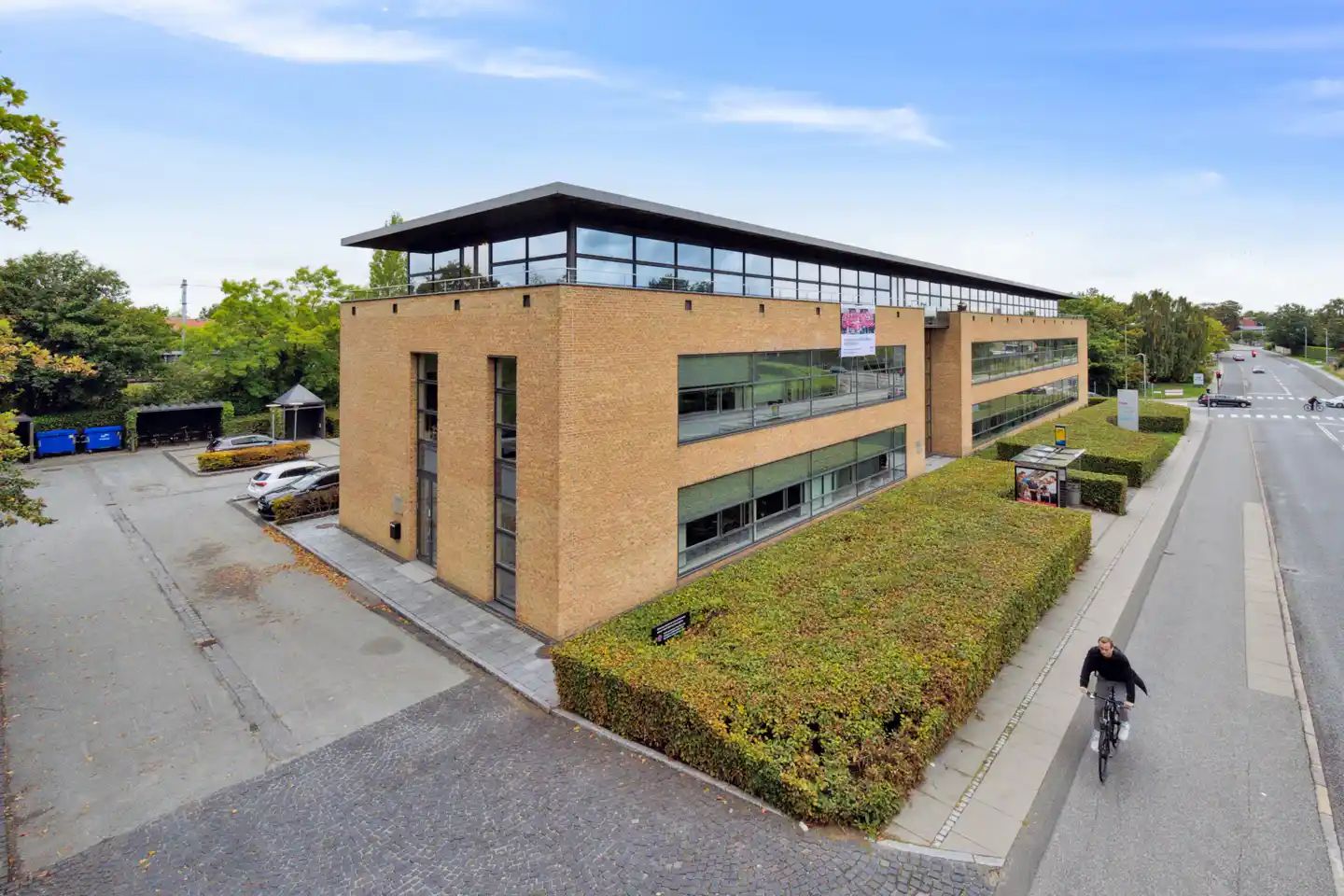 Et moderne og luksuriøst kontorbyggeri med glasvinduer og terrasser, beliggende på en travl vej med grønne buske og