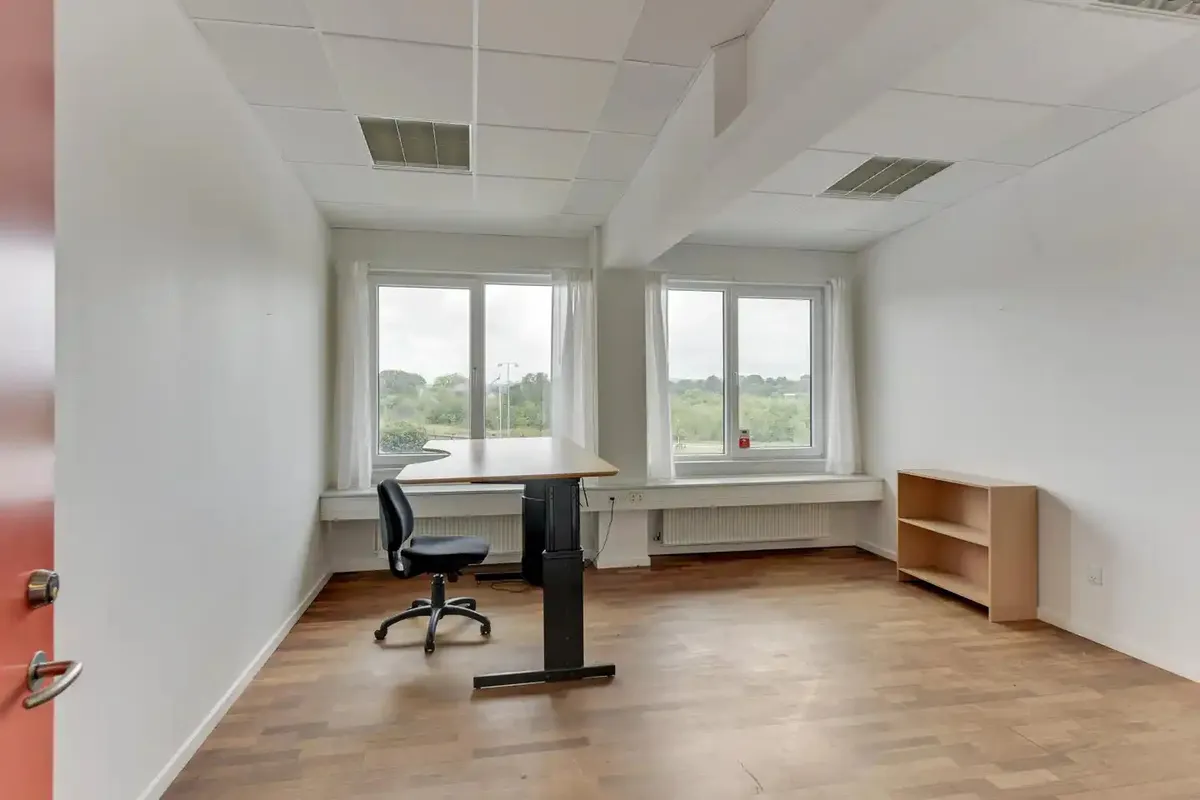 Kontorrum med to vinduer, højt loft, lysegule vægge, tomt skrivebord, høj stol og lille simpel træhylde