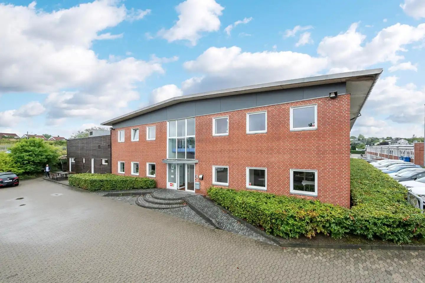 Et moderne kontorlokaler med røde mursten og glasvinduer ligger i en grøn parkeringsplads med en række parkerede biler og en