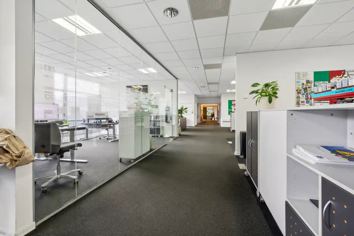 Moderne og funktionel kontor med høje glasvægge, møbler og grønne planter på højre side, trægulve og højloftet