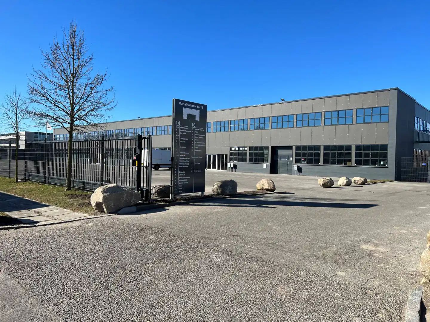 Store bygning med glasvinduer og garagedør. Plads med store sten og træer i baggrunden.