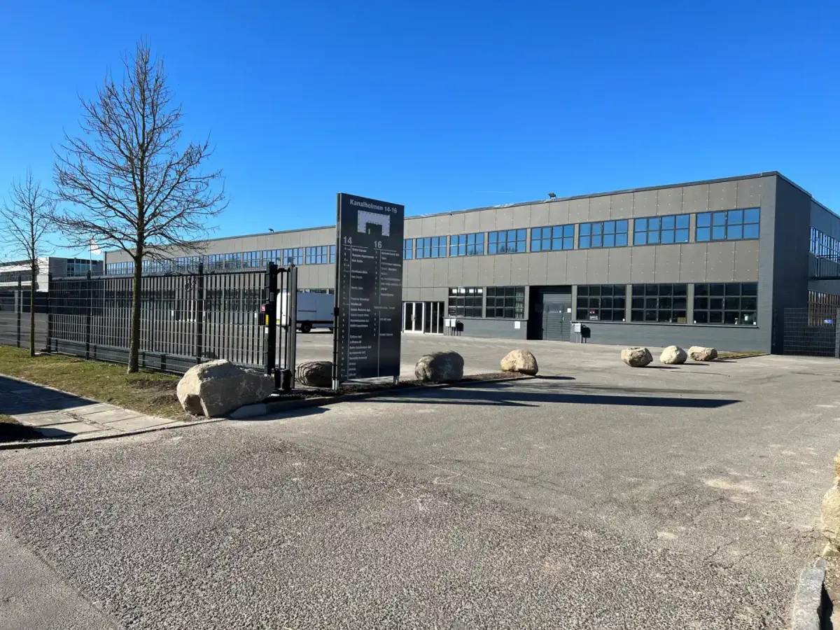 Store bygning med glasvinduer og garagedør. Plads med store sten og træer i baggrunden.