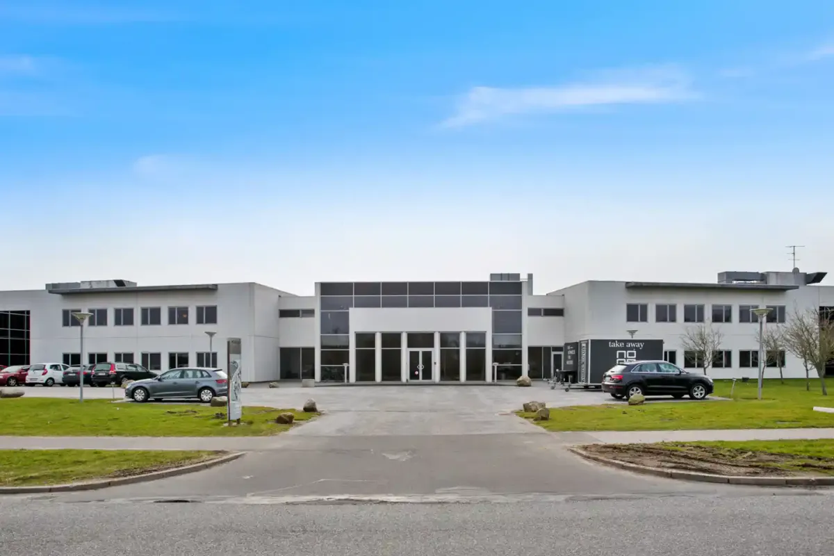 Moderne kontorbygning med glasvinduer og skrifttræk, parkeringsplads og grønne anlæg foran