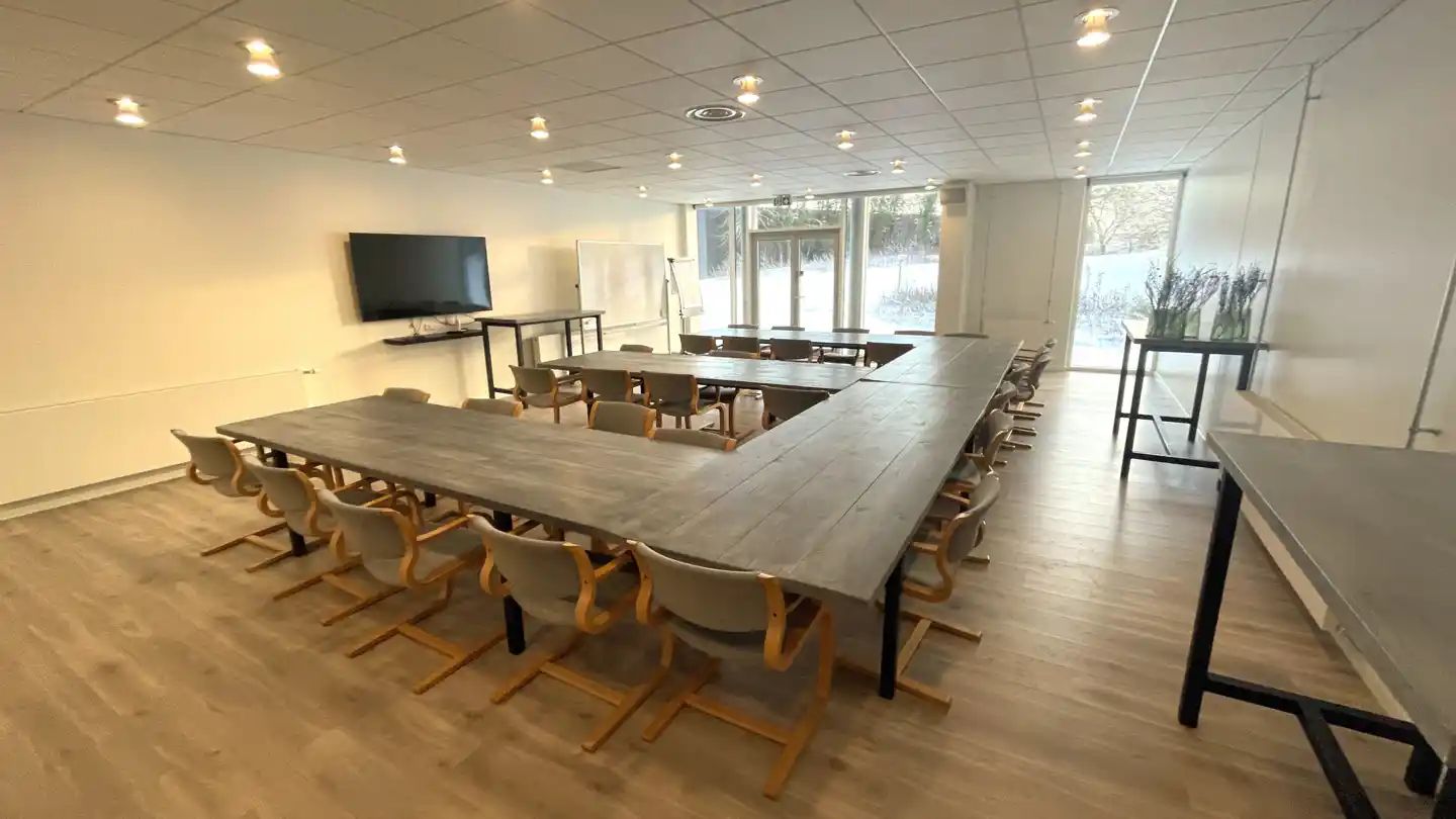 To lange træborde i mødelokale med moderne stolpe møbler og store vinduer med udsigt til skov