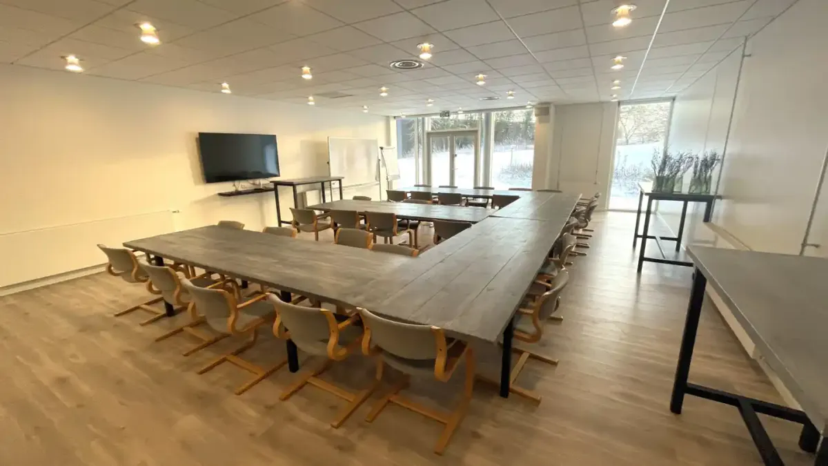 To lange træborde i mødelokale med moderne stolpe møbler og store vinduer med udsigt til skov