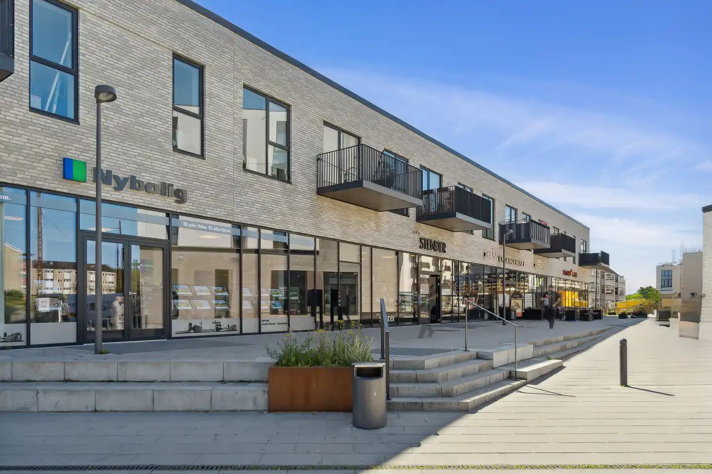 Bygning med facade af lyse mursten og glasvinduer, indgangen markeret med logoet Nybolig og vinduesreklamer