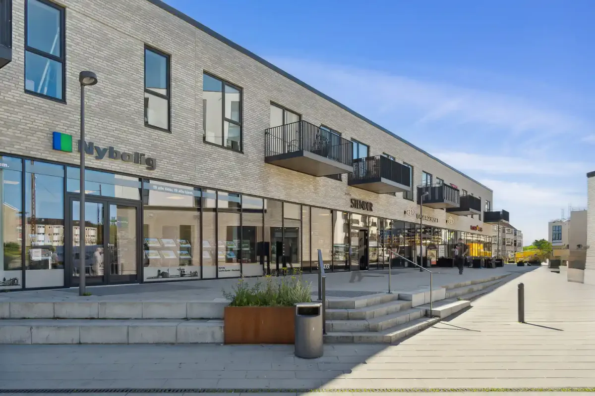 Bygning med facade af lyse mursten og glasvinduer, indgangen markeret med logoet Nybolig og vinduesreklamer