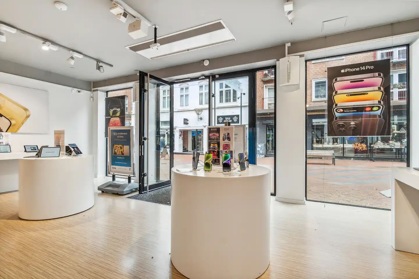 En forretning med hvide rundede borde og iPhone-produkter. En iPhone 14 Pro reklame i vinduet og en banner med teksten 10%