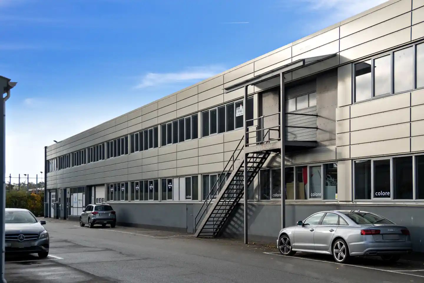 To biler er parkeret udenfor en stor bygning med glasruder og et trappeværk til siden