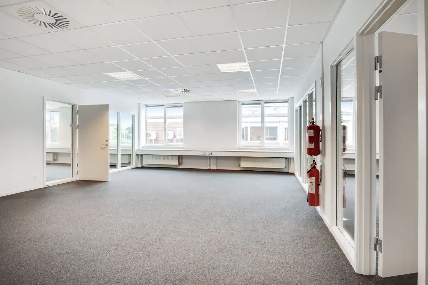 En tom kontorlokal med hvide vægge, gulvtæppe og vinduer. To brandvogne er monteret på væggen. Loftet er hvidt med ventiler