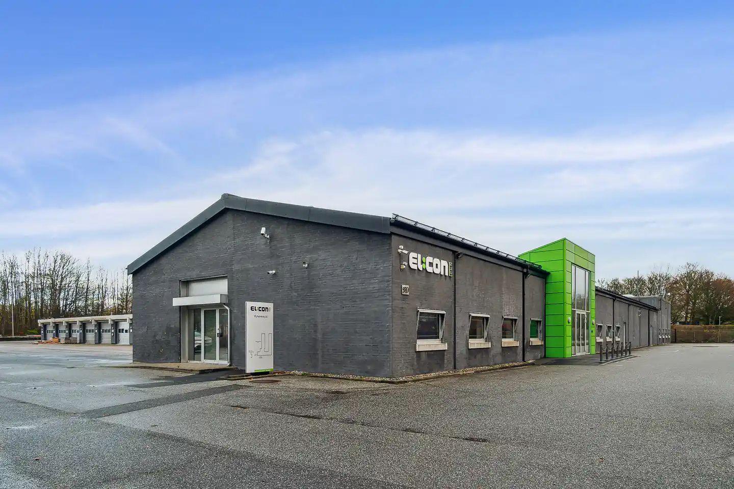 Elcon bygning med grøn facade og glasdøre. Foran er to garageindkørsler og parkeringsplads.