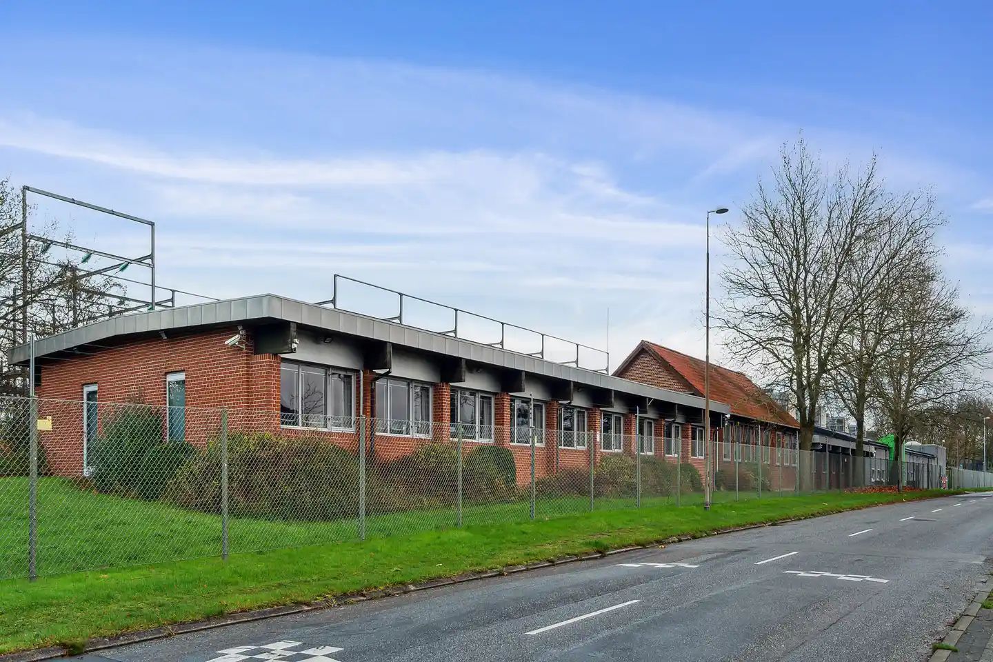 En fabriksbygning med røde mursten og store ruder langs en tom vej med hvide markeringer og en lav græsplæne med buske og et