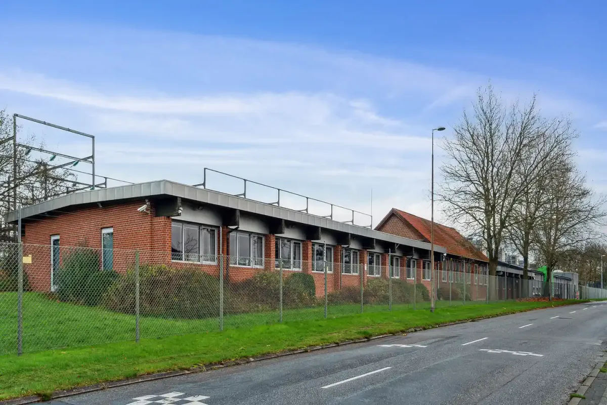 En fabriksbygning med røde mursten og store ruder langs en tom vej med hvide markeringer og en lav græsplæne med buske og et