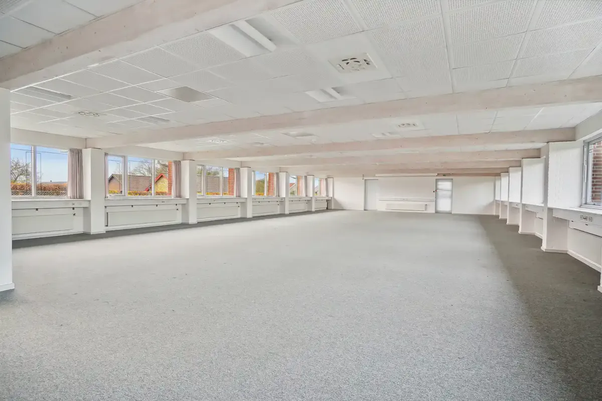 Rummelig og mørk kontorlokaler med hvide lofter og gulve samt flere vinduer mod gårdsplads og træer