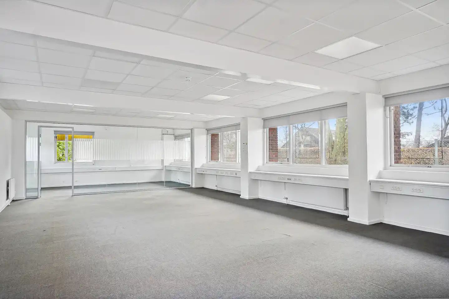 Et stort, ubeboet rum med hvide vægge, gulv og loft samt store vinduer og en glasdør i den ene ende