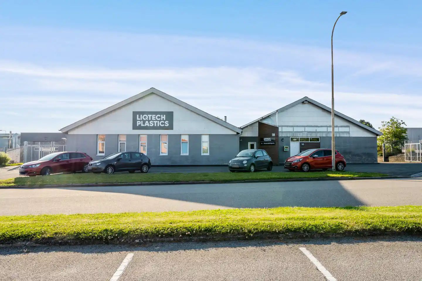 Første og anden etage af et byggeri med skiltet Liqtech Plastics. Parkerede biler foran bygningen og en lysstolpe på græsset.