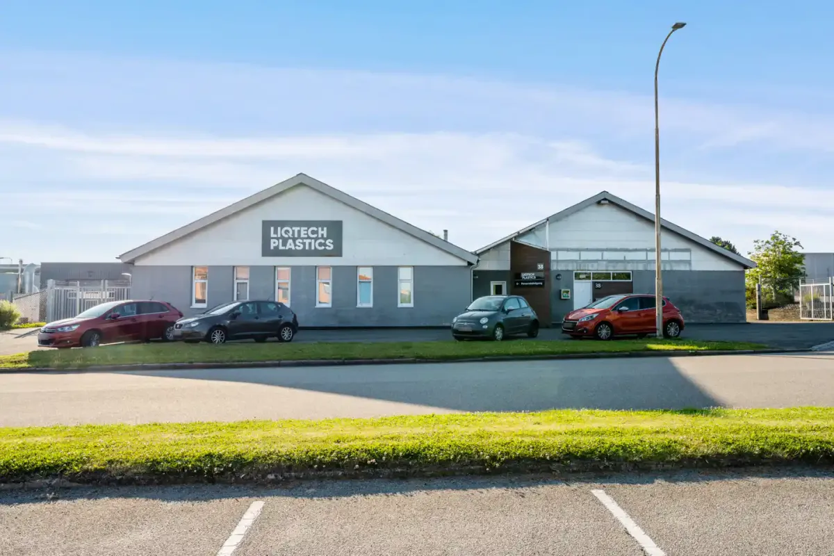 Første og anden etage af et byggeri med skiltet Liqtech Plastics. Parkerede biler foran bygningen og en lysstolpe på græsset.