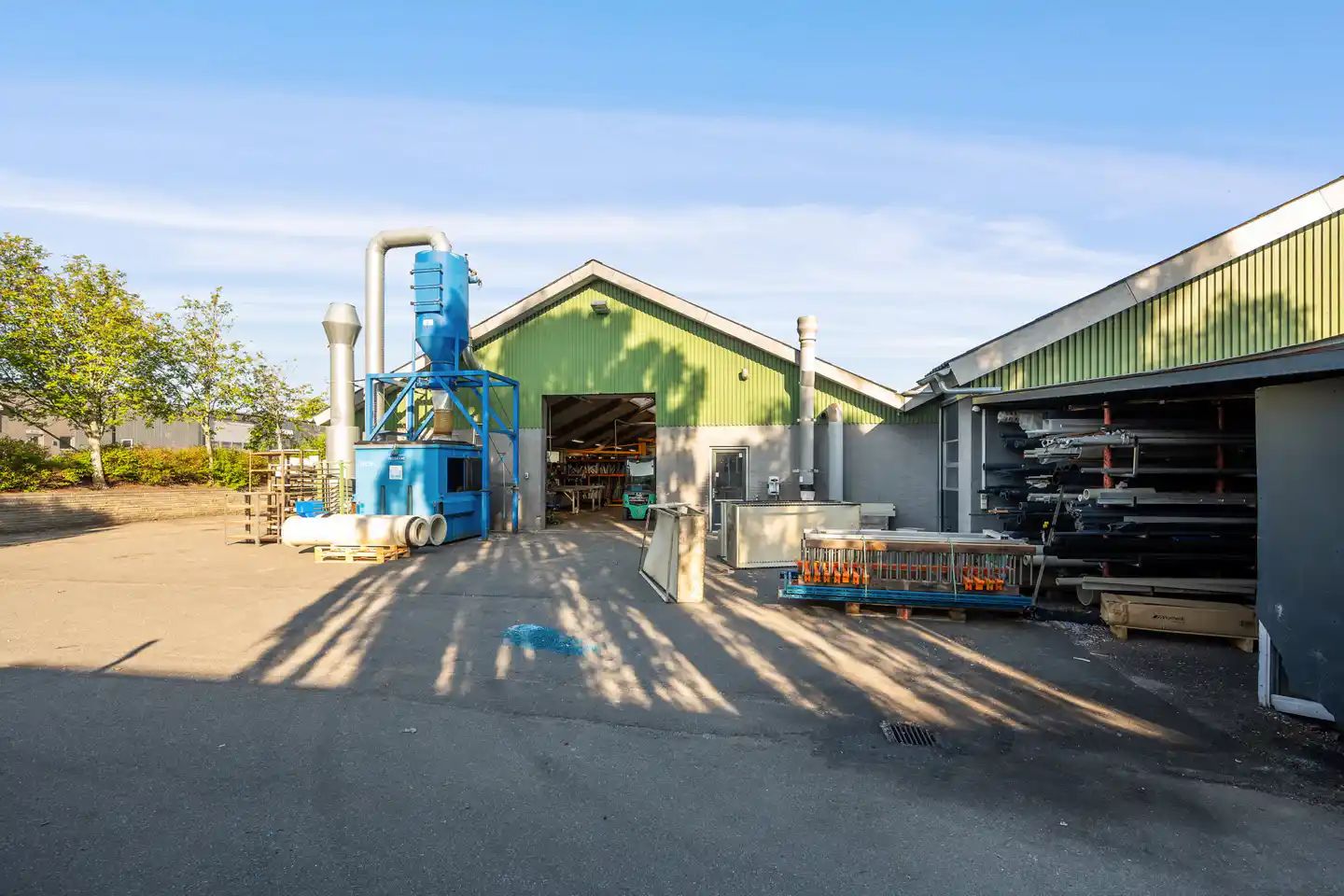 To bygninger med grønne lofter og åbne døre med indre lager og diverse materialer og maskiner og en stor blå og hvid