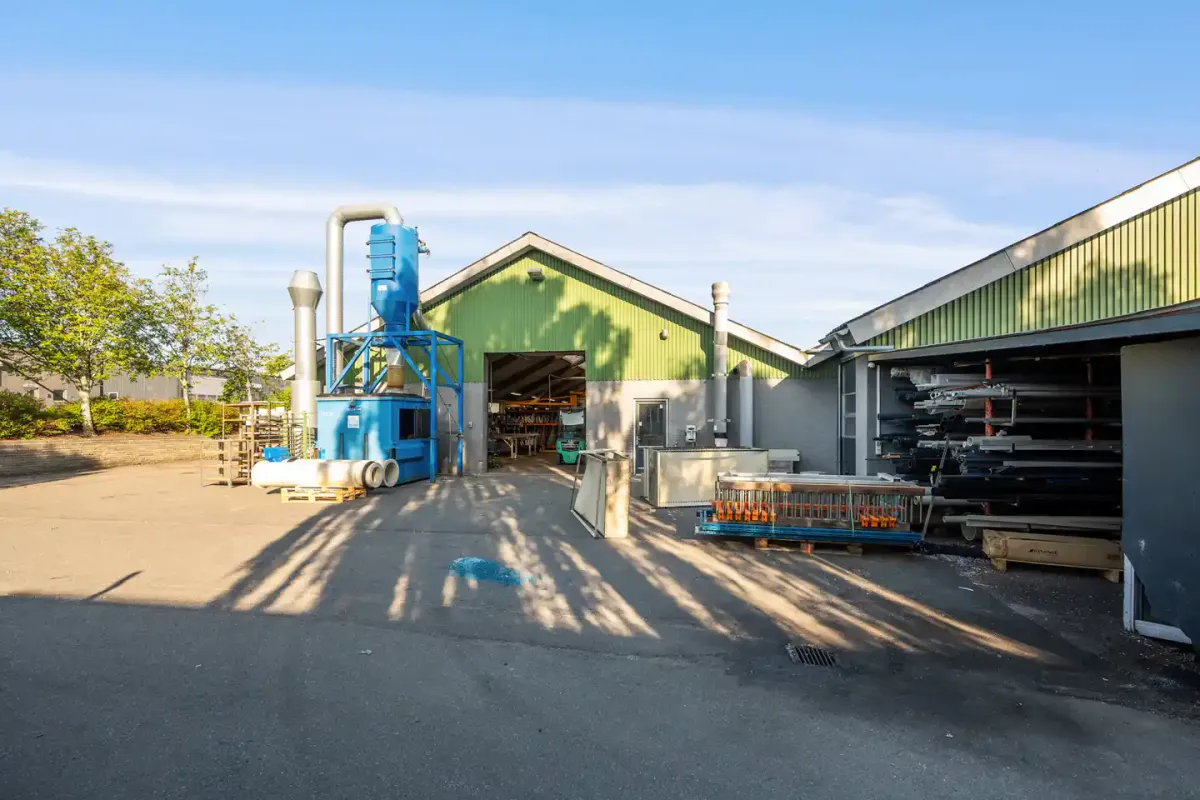 To bygninger med grønne lofter og åbne døre med indre lager og diverse materialer og maskiner og en stor blå og hvid