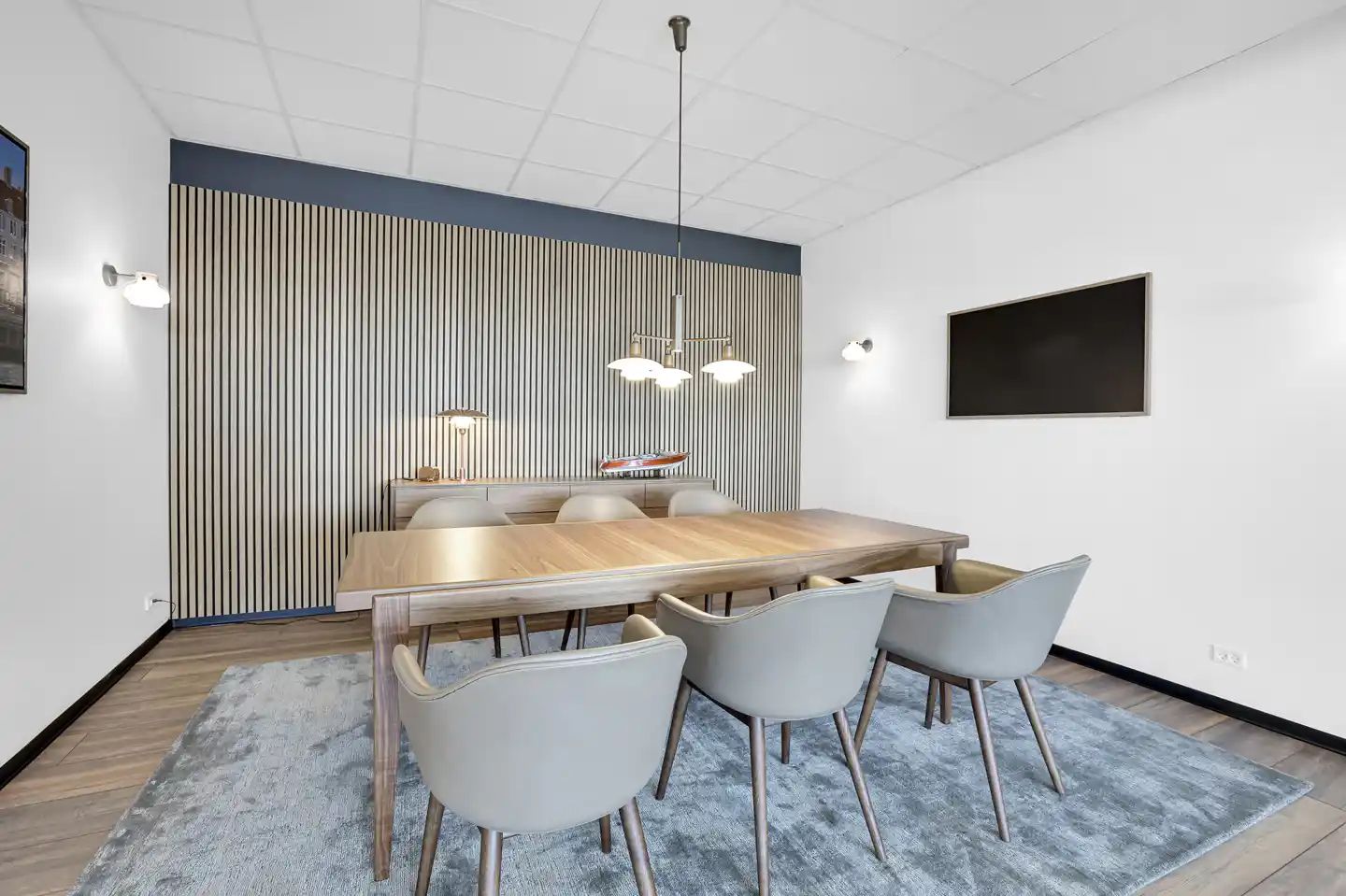 En luksuriøs mødelokale med lyse træborde og stole, hængende lysekroner, og moderne indretning med en flatscreen-tv
