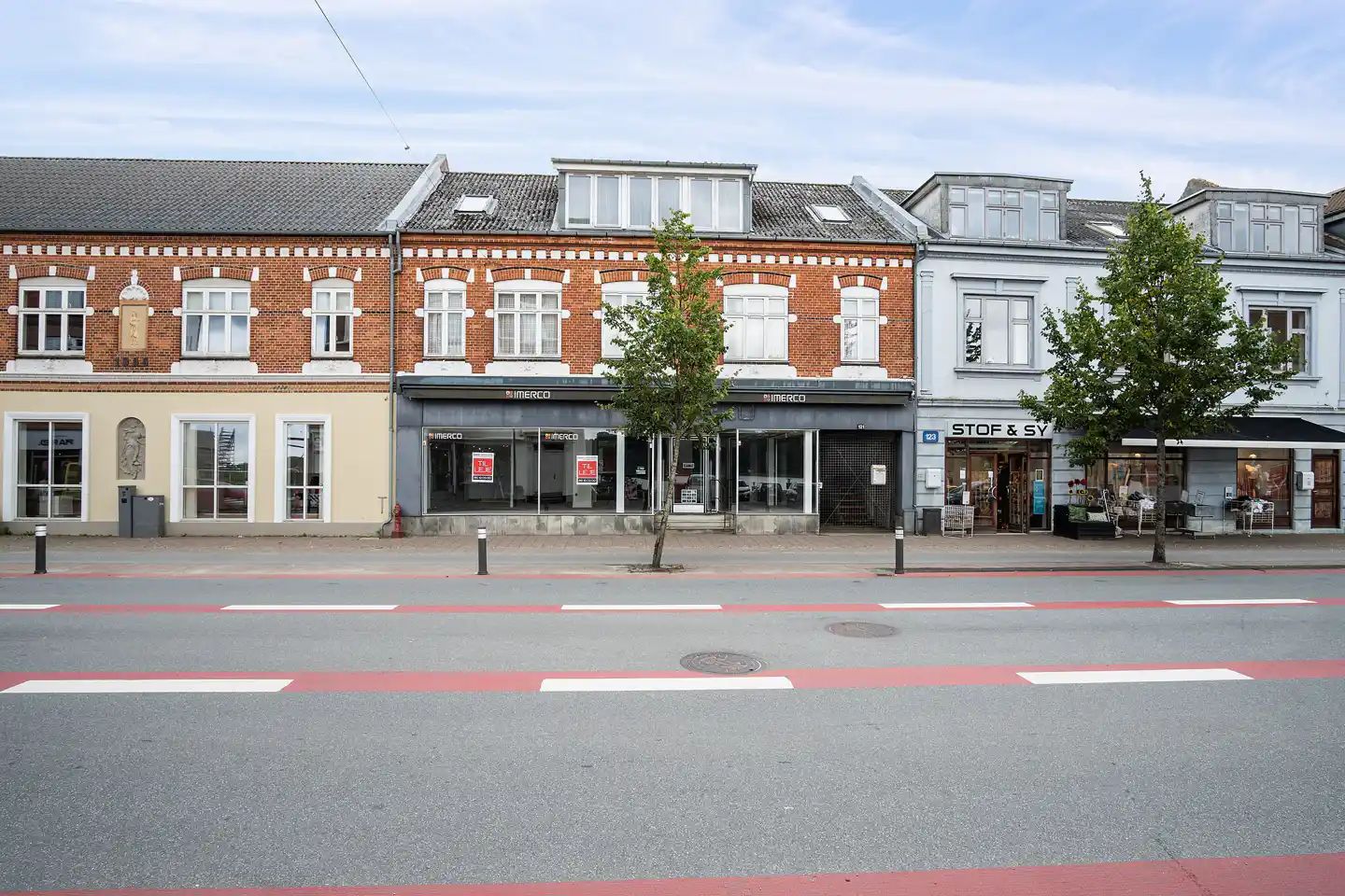 Gade med tre bygninger med store vinduespartier og en stor træstamme i forgrunden