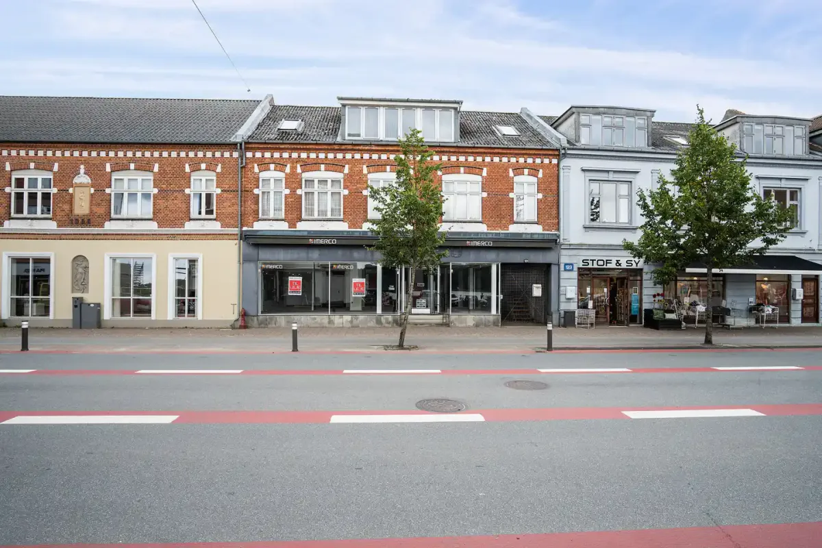 Gade med tre bygninger med store vinduespartier og en stor træstamme i forgrunden