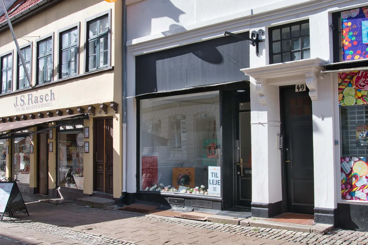 Butik med udsalgsvindu og indgangsdør, der læser Til Leje. I udsalgsvinduet er der blomster og en skilt med teksten Til