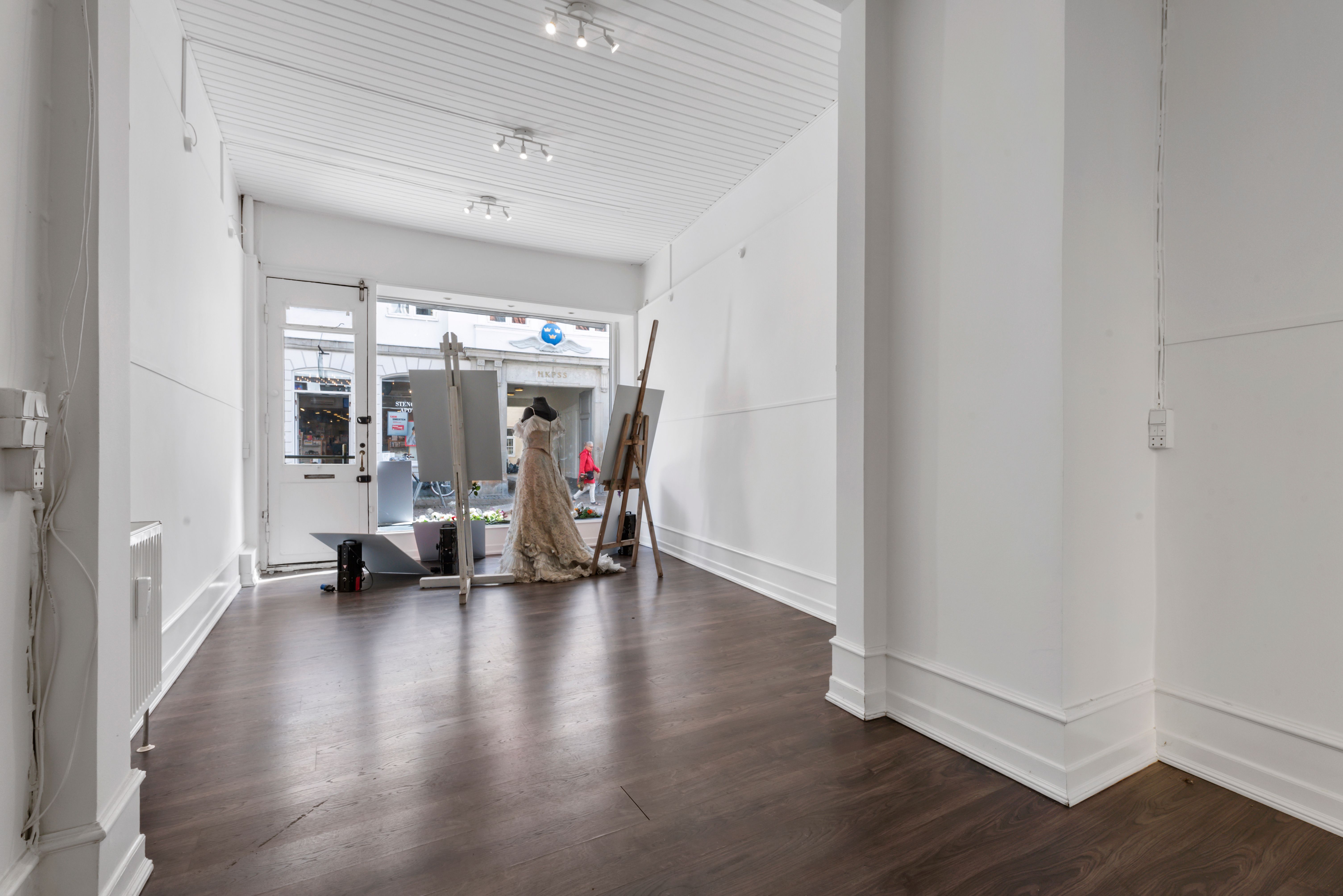 Lukket butik med hvide vægge og gulve, hvor et ægte brudekjole er opstillet med to malerstav. Der er en person udenfor, og