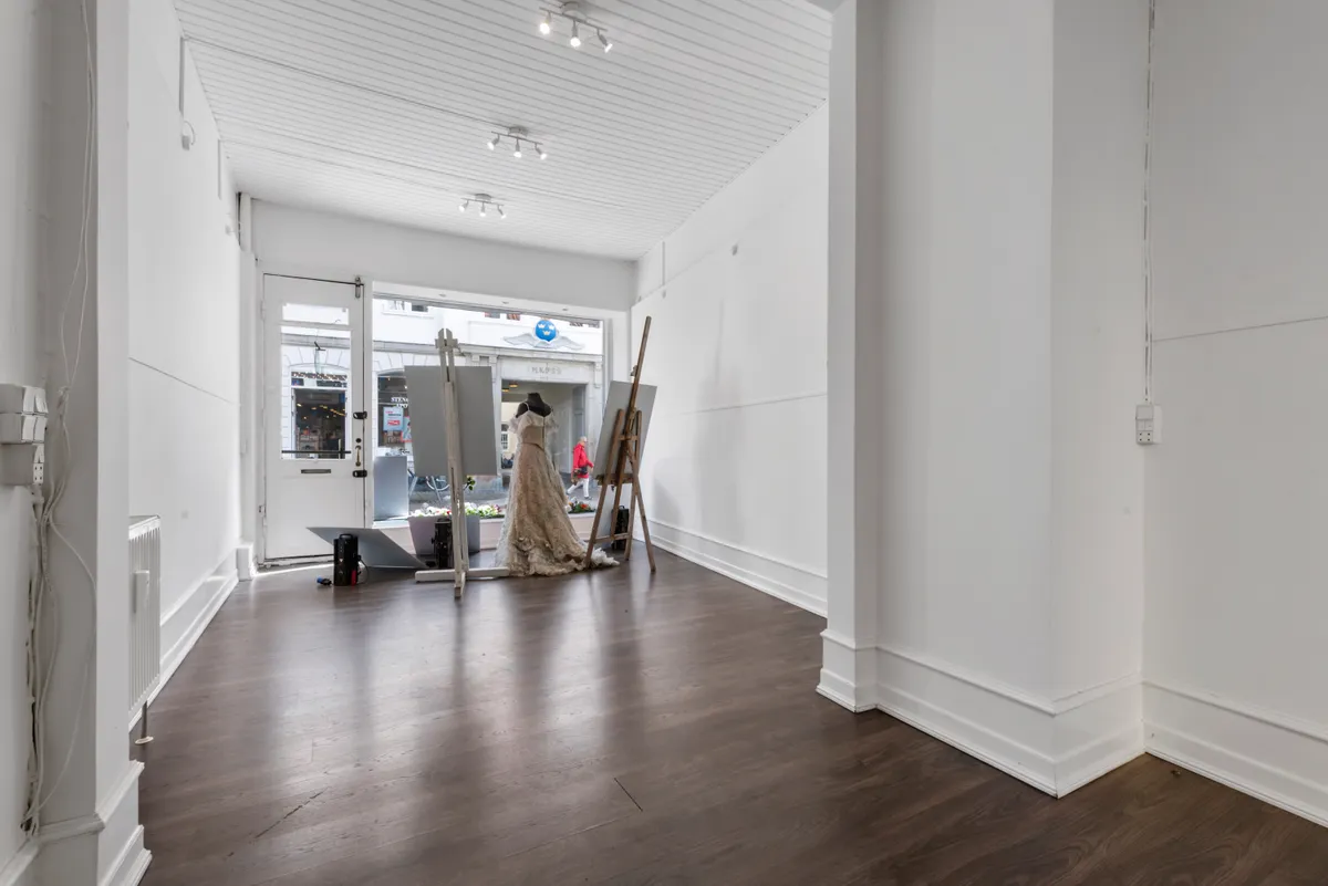 Lukket butik med hvide vægge og gulve, hvor et ægte brudekjole er opstillet med to malerstav. Der er en person udenfor, og