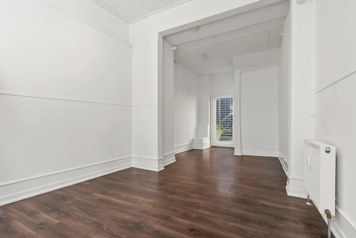 En stor hvid stue med højt loft og mørk trægulv. Der er en hvid radiator og et hvidt vindue med metalgitter i baggrunden.