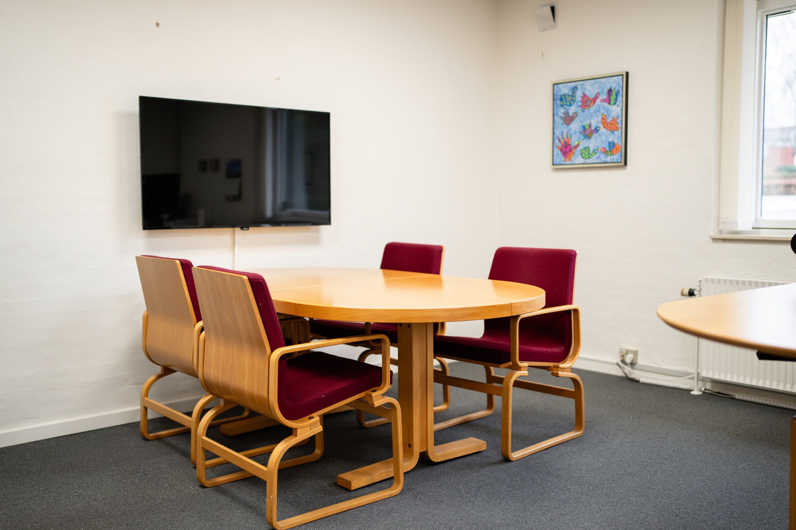 Rundt kontorbord med fire røde stole og en tv-skærm samt et billede i lysegrå væg