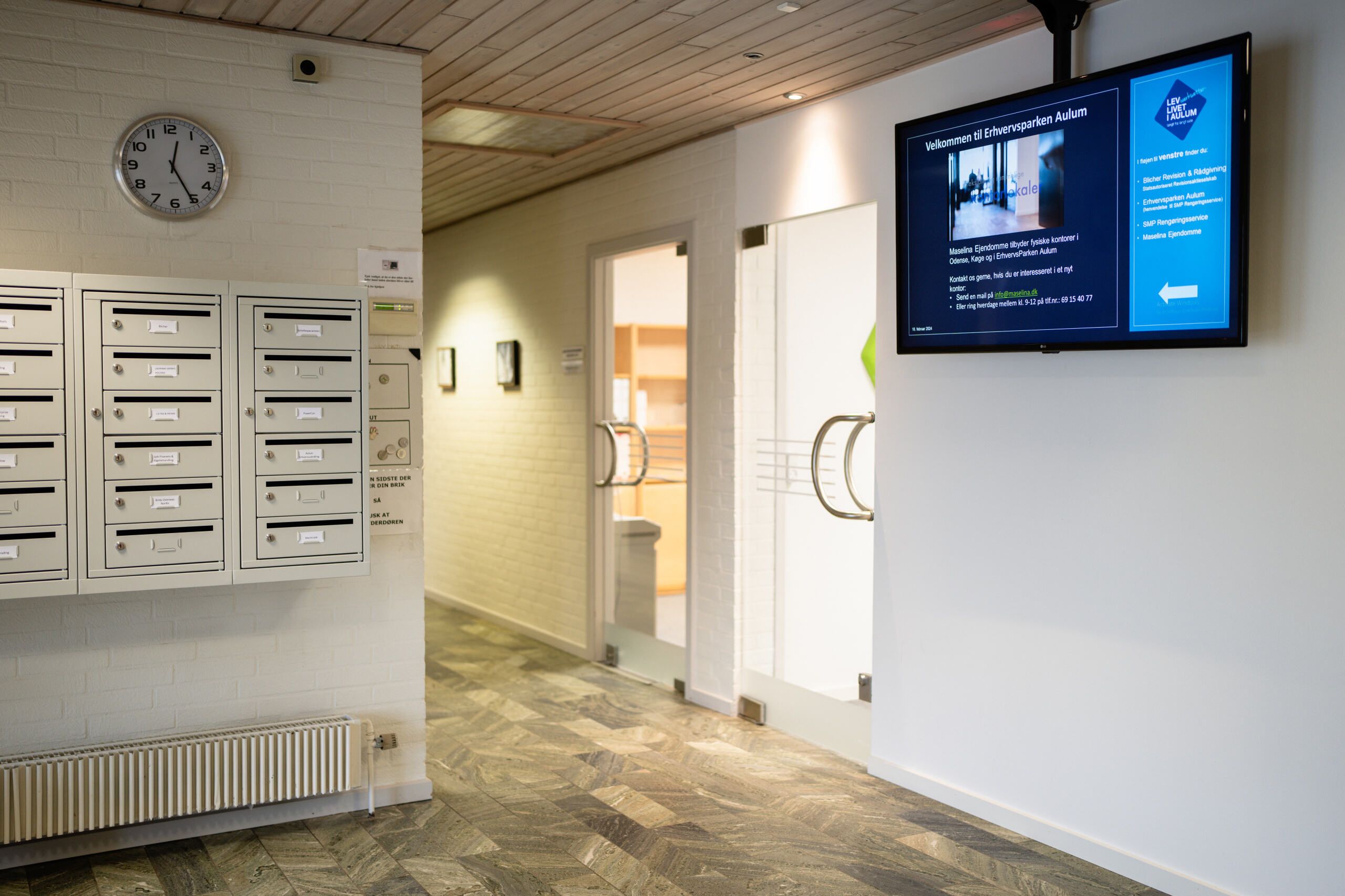 Et moderne kontor med lyse vægge, en stor skærm, hængende ur, åbne døre og flere postkasser og radiatorer.