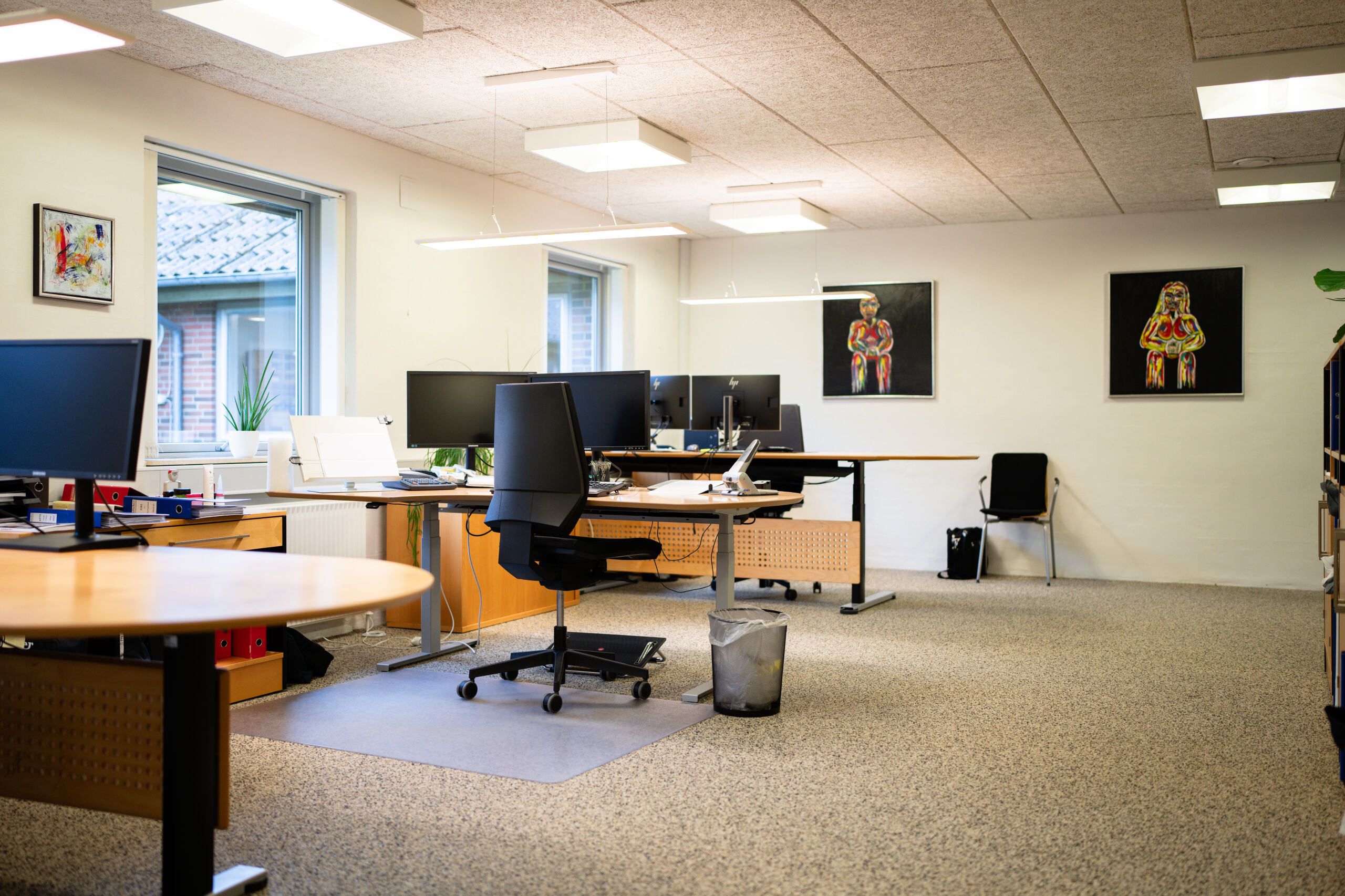 Kontor med flere borde og stole, computere og skrivebordsplanter. Billeder hænger på væggen og der er lysekroner og lyse