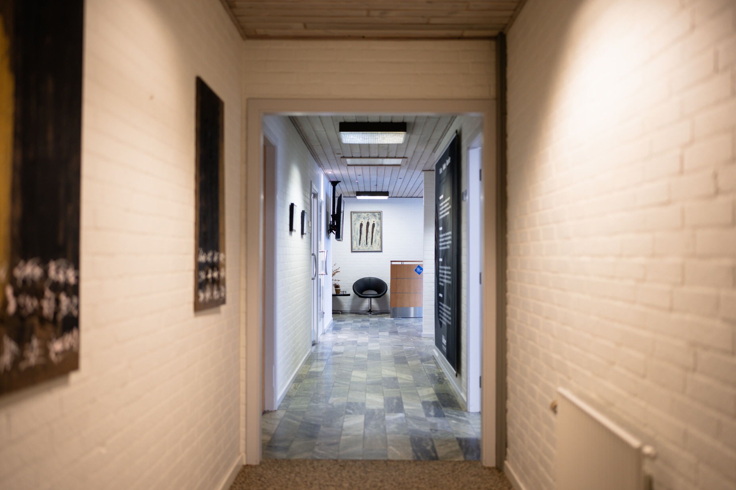 En lang, lys hall med hvide vægge og mønstrede gulve, to tv'er og en stol samt et bord med plante.