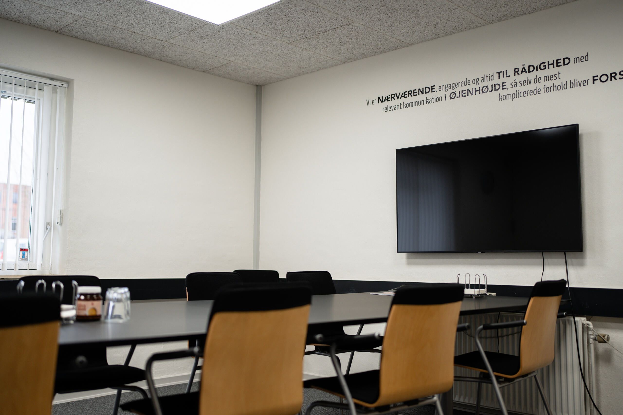 Kontorrum med bord, stole og tv. Der er en mørk farvet tv-skærm monteret på væggen. Der er tekst på væggen.