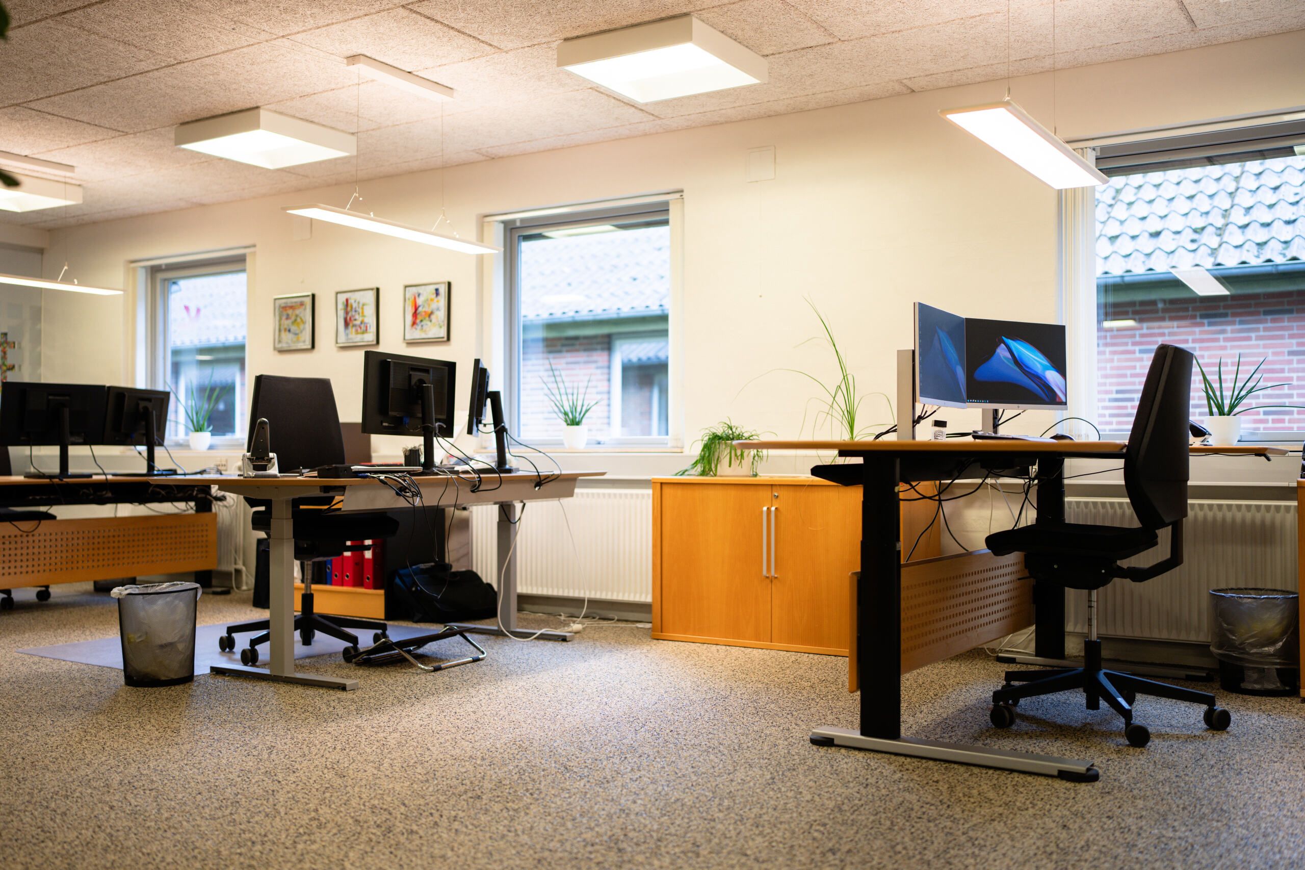 Kontor med lyse loftslampe, skrivebord, stol, plante, og vinduer med udsigt til en bygning med tegltag.