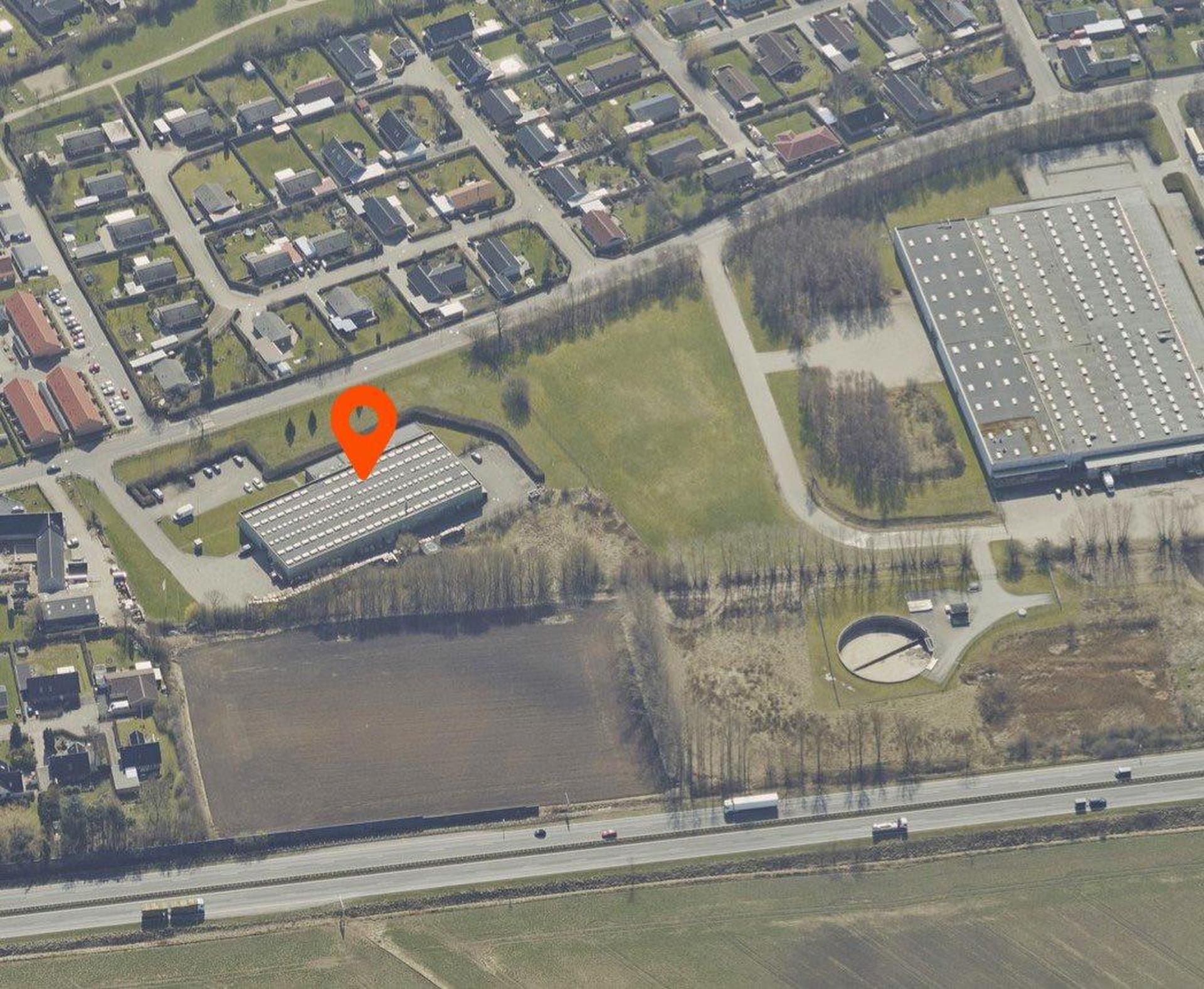 Flyfoto af en stor erhvervsejendom med parkeringsplads, beliggende ved en hovedvej med flere lastbiler, samt