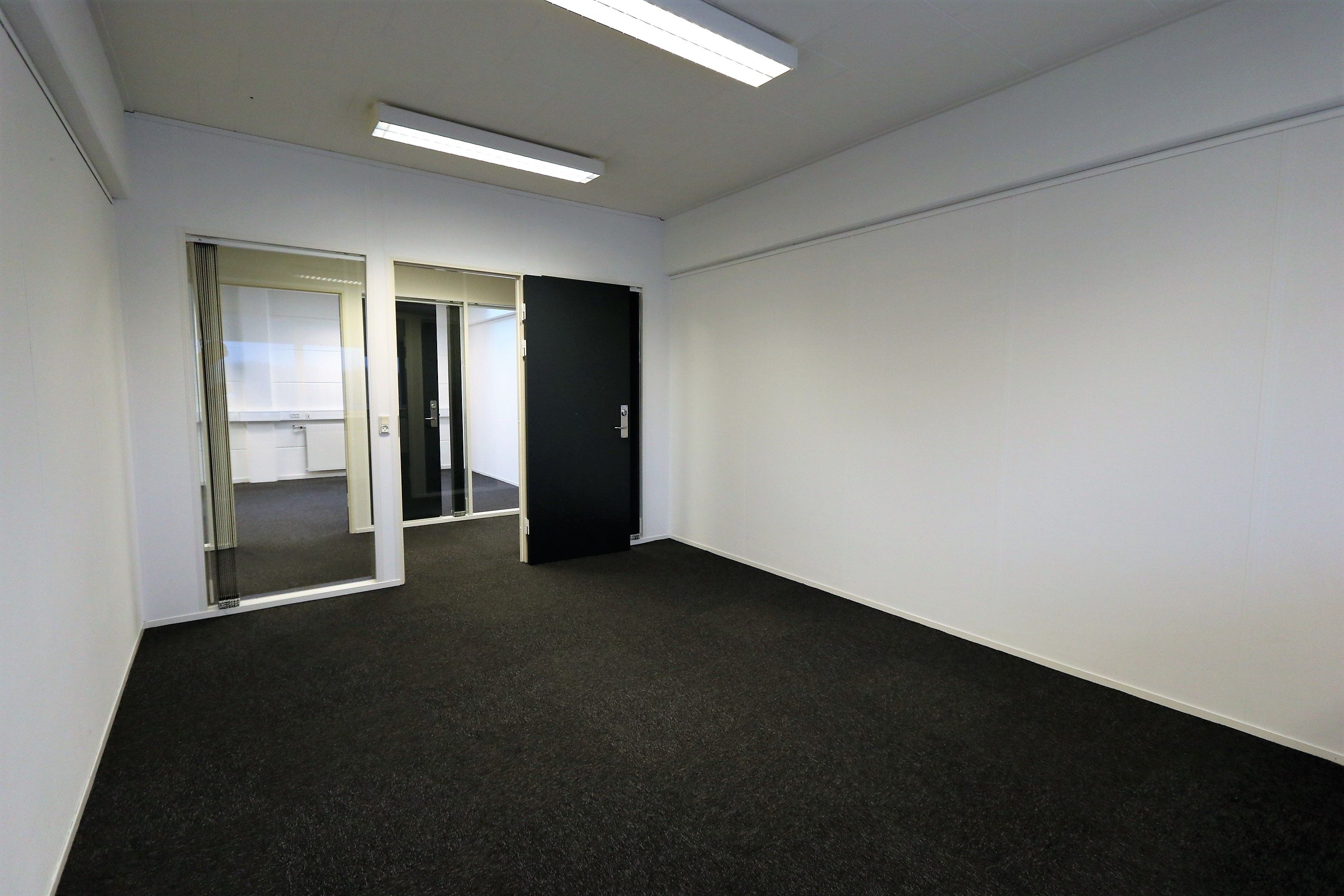 Lukket og indflytningsklart kontorlokale med glasdøre og sort trægulv. Væggene er hvide og der er en lysekron på loftet.