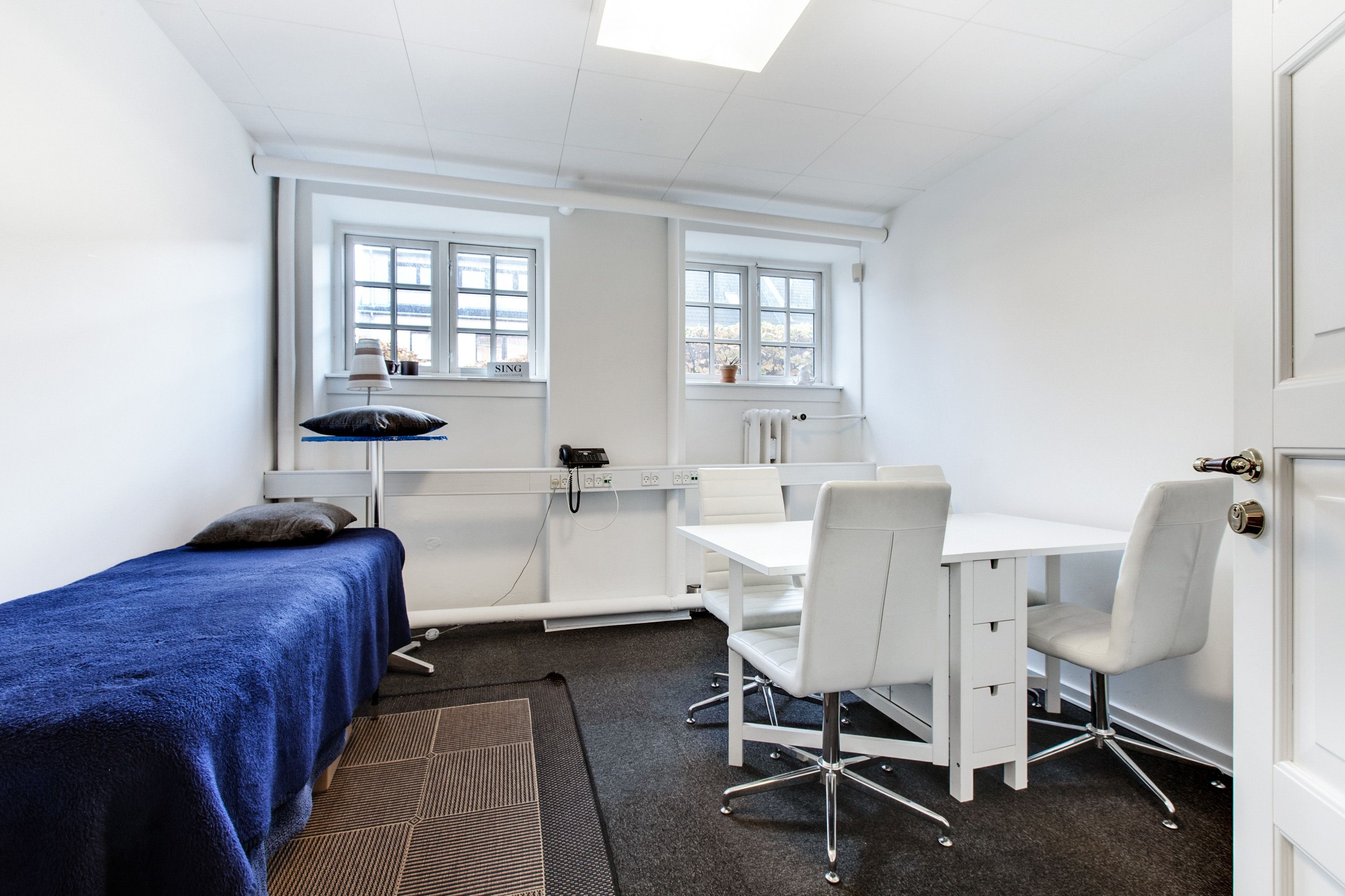 En hvid stue med en hvid bord og fire hvide stole, et sovesofa med blå tæppe, et skrivebord med telefon og lampe og et