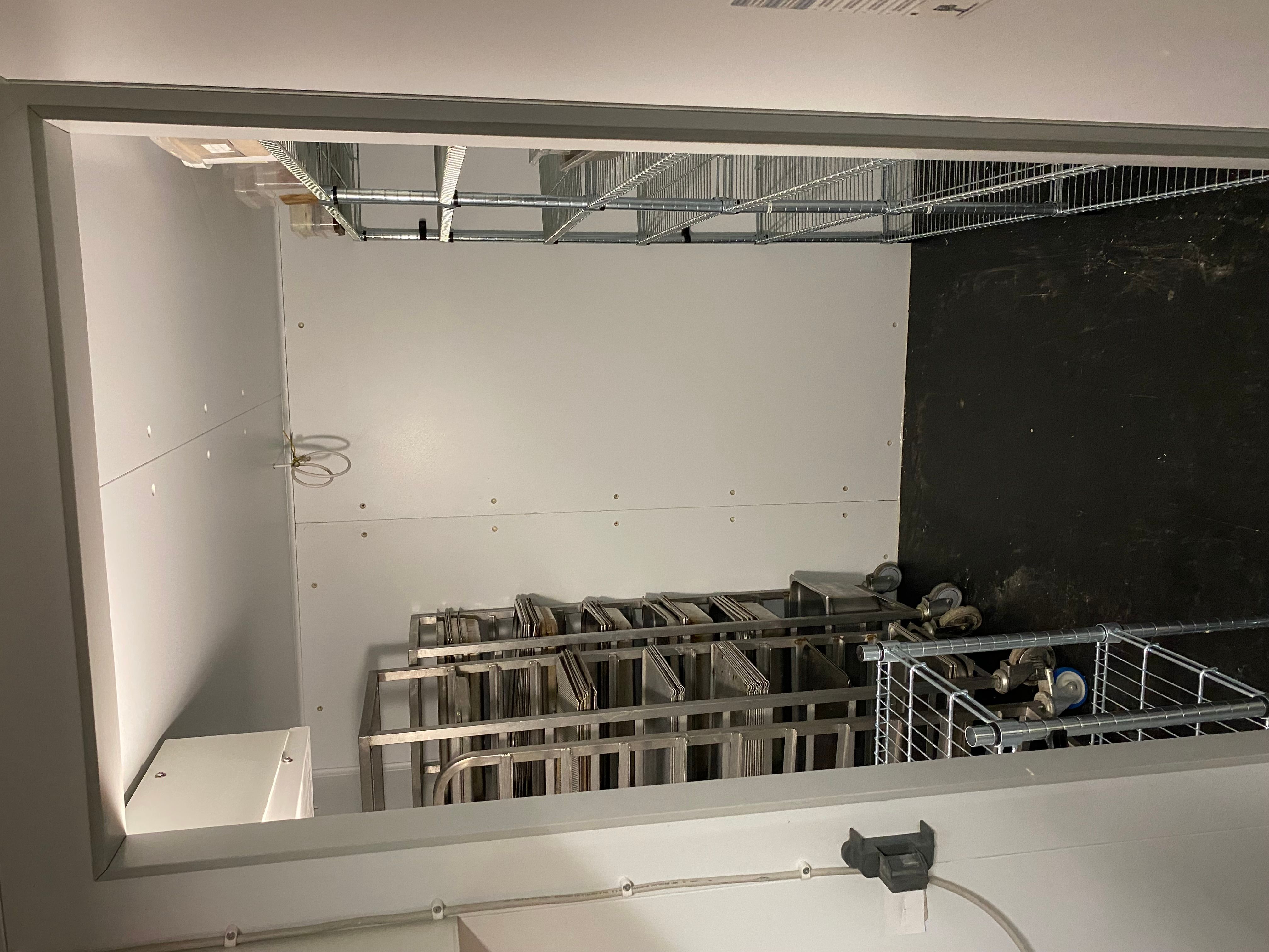 Et stort produktionskøkken med et stort kølekammer og flere stålhylder. Stålhylderne er fulde med fødevarer og plastposer.